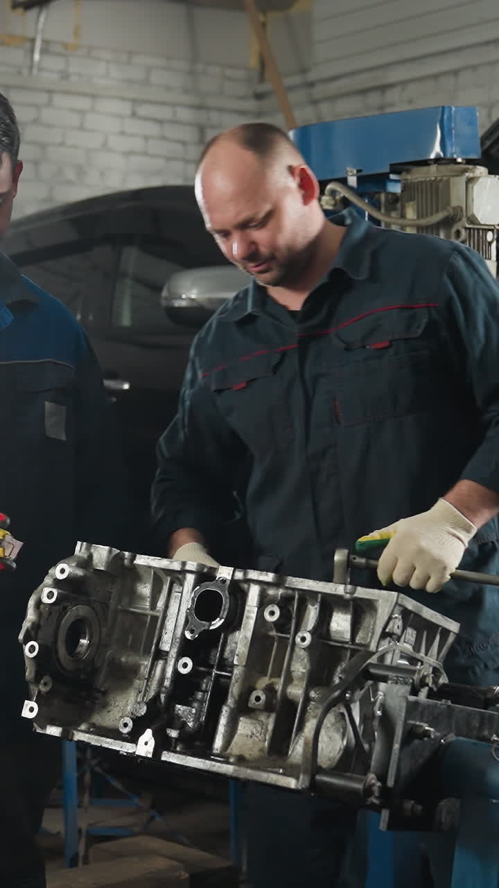 dos mecánicos en uniformes azules, uno pasando una herramienta a otro mientras trabaja en un motor en un taller bien equipado, el fondo muestra un coche y varias herramientas mecánicas