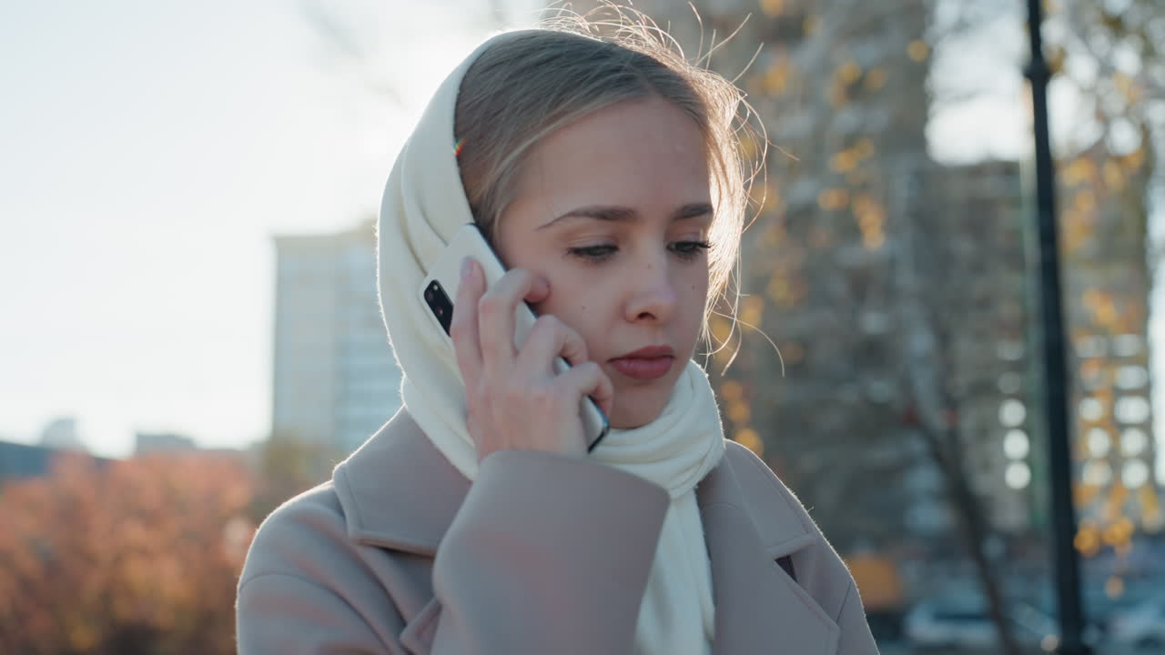 Caucasian woman student on phone, autumn park backlight, beige coat and cream scarf, checking mobile then calling, concerned expression, city buildings and soft sunlight, chilly breath, cinematic