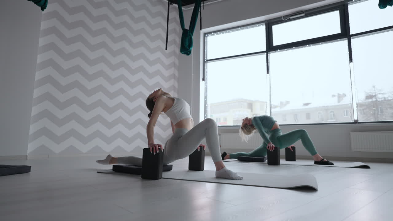 Group of young women stretching in gym with windows. Two attractive sport girls work out nauka asana boat yoga pose on black mat in fitness class. Two women stretching legs together in yoga studio