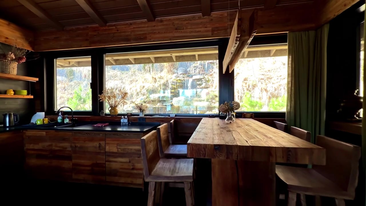 Cucina e zona pranzo di una baita in legno con grandi finestre che si affacciano su una cascata rocciosa.