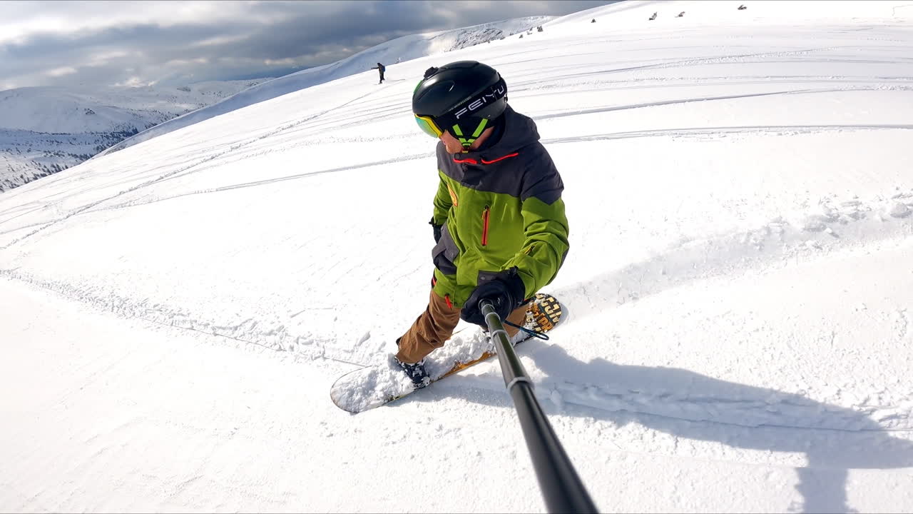 Male snowboarder in special outfit and protective glasses and helmet goes down the snowy hill. Man taking selfie of his ride and beautiful mountains at backdrop.