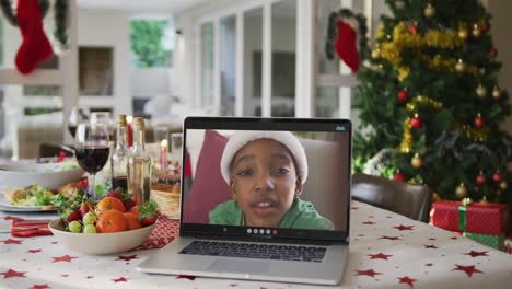 feliz niño afroamericano en el sombrero de santa en la computadora portátil acostado en la mesa de navidad