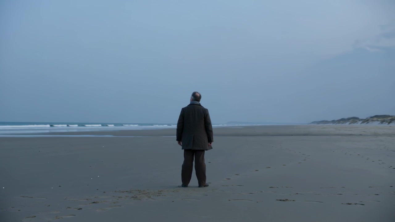 Persona mayor de pie con abrigo oscuro y pantalones, observando las olas del océano en la playa, después de caminar hasta la orilla