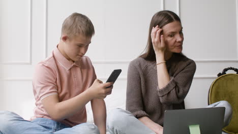 Junge-Mit-Down-Syndrom-Und-Seine-Mutter-Mit-Smartphone-Und-Laptop-Sitzen-Auf-Dem-Bett-Im-Schlafzimmer-Zu-Hause