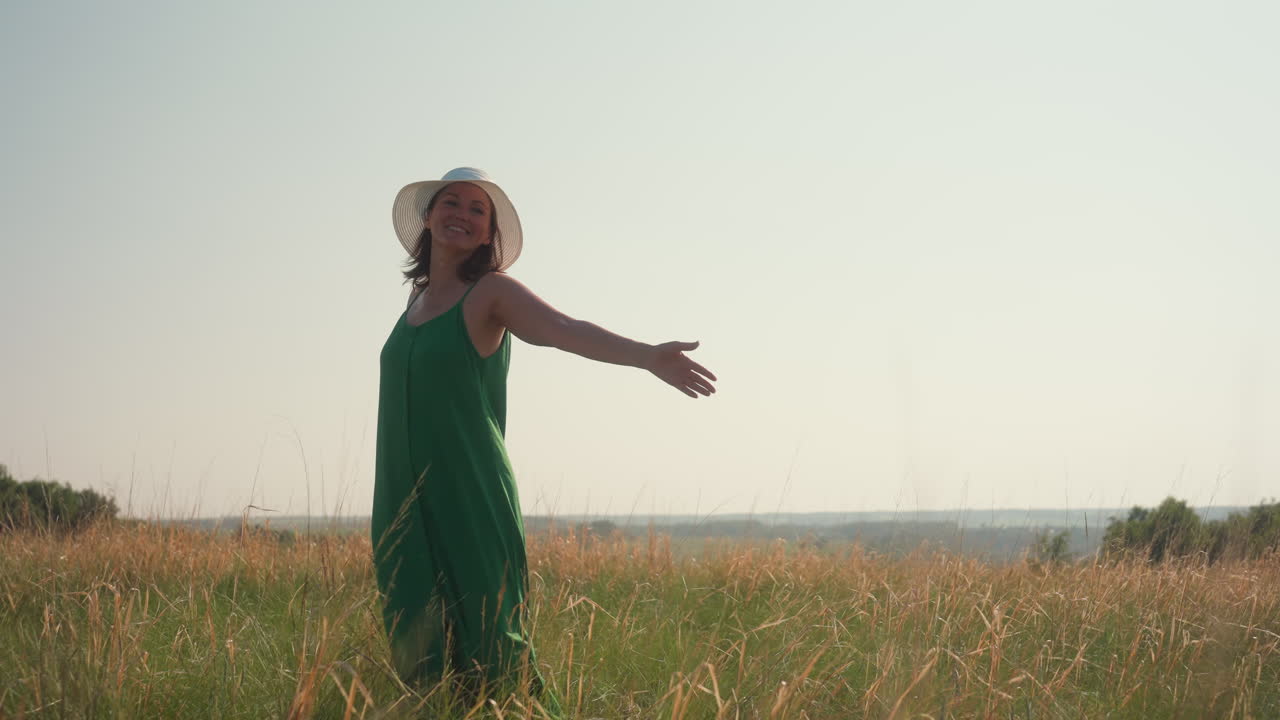 woman in wide hat dances joyfully with outstretched arms in tall grassy field under soft sky, wearing flowing green dress, smiling freely in nature during warm day surrounded by gentle breeze