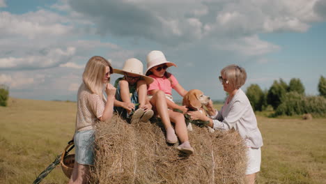 family enjoys outdoor fun on sunny day, children wearing straw hats, grandma feeds dog on haystack, woman adjusting sunglasses, countryside scenery, rolling hills, trees in background