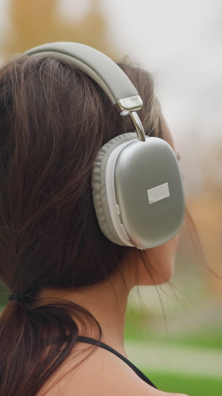Young lady standing and placing headphones on her head with a blurred background of a serene outdoor environment, she appears calm and focused as she adjusts her headphones for a listening experience