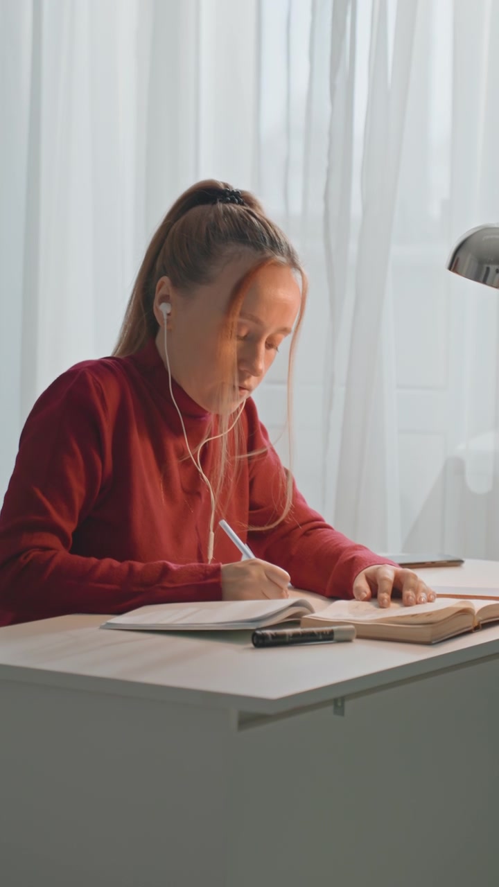 Young woman in red sweater studies at desk with open book. Woman writes notes while reading from text occasionally pausing. Bright room features white curtains silver lamp