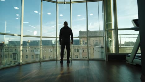 back view of a man in the gym looks out the panoramic window at the city view