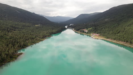 Stunning-Aerial-View-of-a-Green,-Teal,-Turquoise-Long-Lake-with-a-in-Norway,-Reflections-on-the-Calm-Water,-Norwegian-Forest-Road,-Some-Traffic-in-the-Background,-Scandinavia