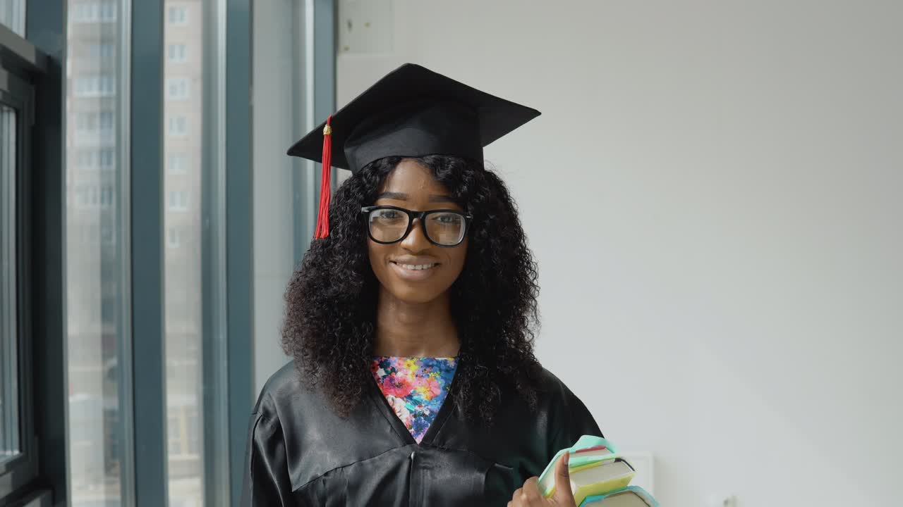 joven graduada universitaria afroamericana de pie con libros de texto en las manos cerca de una gran ventana moderna con marcos negros. estudiante de sexo femenino con una túnica negra, sombrero de maestro cuadrado y gafas
