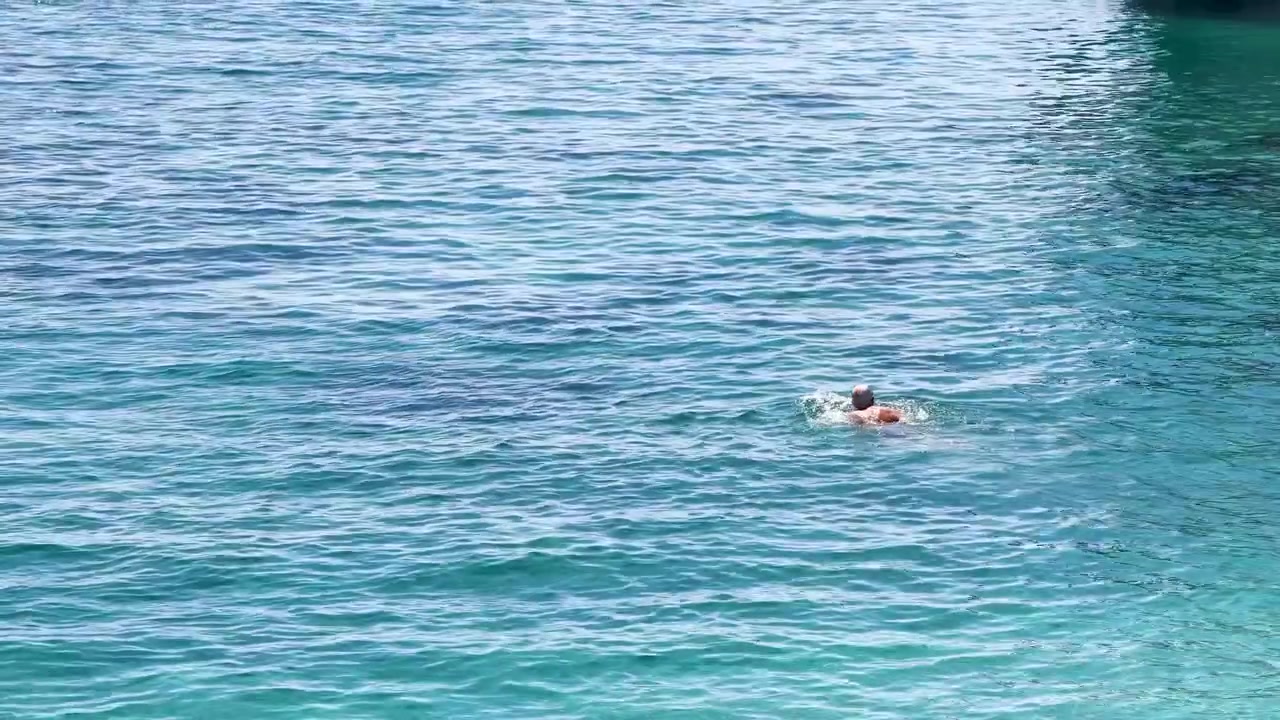 明るい日差しの中、透明なターコイズブルーの地中海の海を泳ぐ人物の広角映像。
