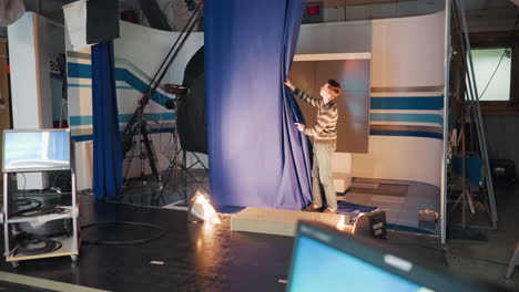 studio stagehand in striped sweater pulls large blue curtain across set preparing tv shoot, floodlights illuminating camera rigs cables visible around space, capturing behind scenes setup mood