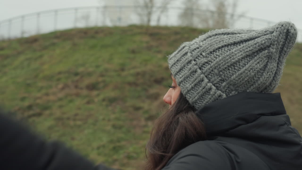 Open arm winter dance on promenade, woman with grey beanie and puffer jacket faces skyline with arms outstretched, overcast sky and railing backdrop, slow expressive movement and reflective mood