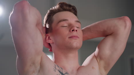 teenager stands with eyes closed and both hands behind head opens his eyes, highlighting toned arms and chest tattoo while expressing confidence and peaceful strength in softly lit indoor setting