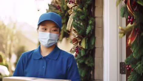 delivery person in blue uniform and face mask brings package to a festively decorated doorway surrounded by greenery and holiday ornaments
