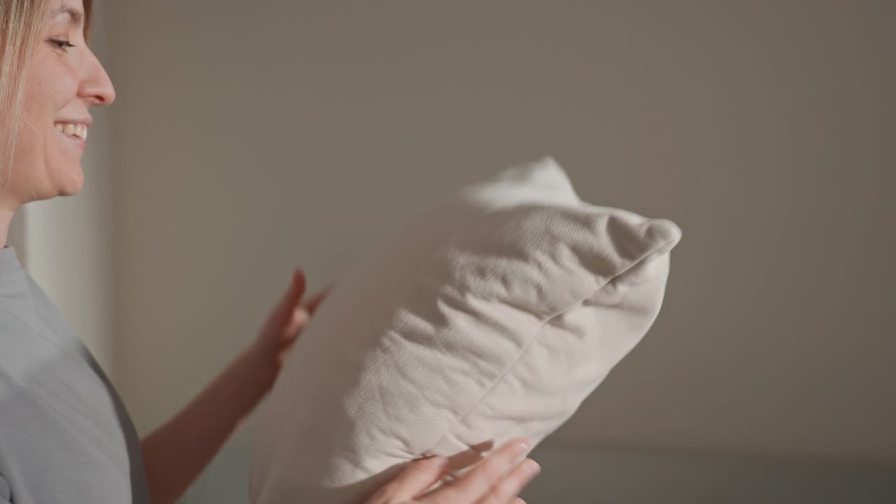 Donna che tiene un cuscino in primo piano accanto a un divano grigio, illuminazione soffusa e interni minimalisti, gesti delicati, atmosfera calma e momento domestico intimo che suggerisce un rituale di ascolto musicale