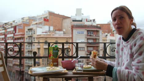 Frau-Lächelt-In-Die-Kamera,-Während-Sie-Im-Pyjama-Auf-Einem-Gemütlichen-Balkon-Zum-Frühstück-Venezolanische-Arepas-Genießt