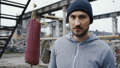 Close-up-view-of-caucasian-man-in-black-beanie-looking-at-the-camera-with-serious-expression-outdoors-an-abandoned-factory-on-a-cloudy-morning