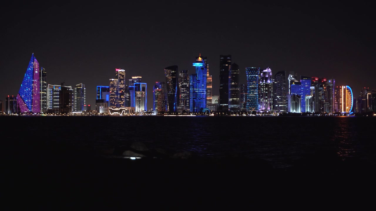 Fantastisk nattutsikt over den opplyste silhuetten av Doha West Bay i Qatar. Med livlige blå og lilla skyskrapere som reflekteres over det mørke vannet i Persiabukta.