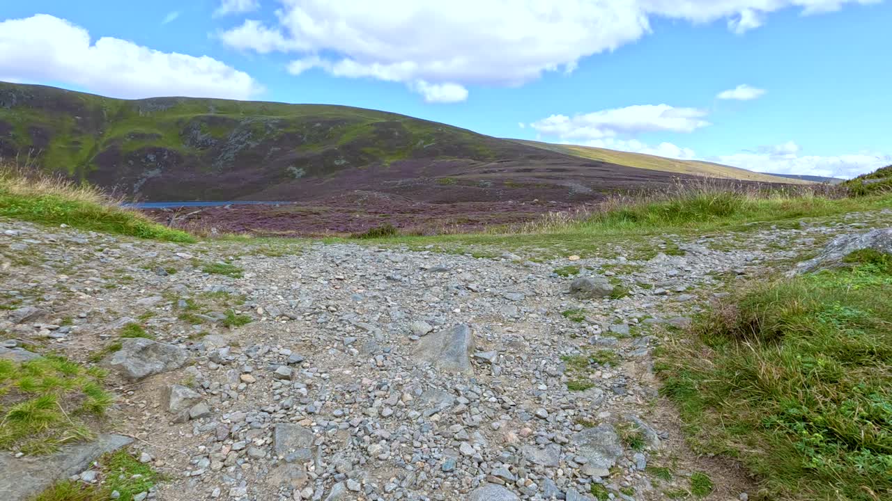 Camera moves along a rocky path through heather-covered hills toward Loch Brandy, under bright daylight with clear blue skies and scattered clouds