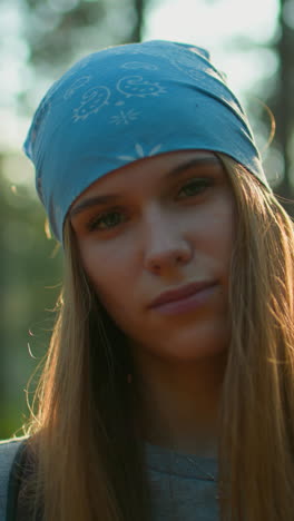 joven con pañuelo azul inclina la cabeza hacia la derecha con una suave sonrisa en el bosque, la luz del sol proyecta un brillo cálido alrededor de su cara, la iluminación natural y el fondo del bosque crean una atmósfera serena