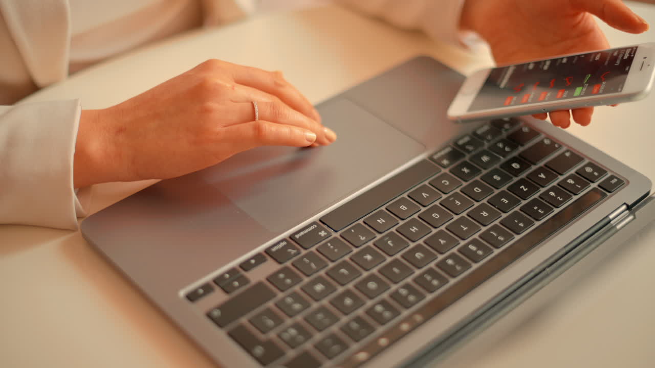 Woman in a white blazer working on a laptop at a table while holding her phone
