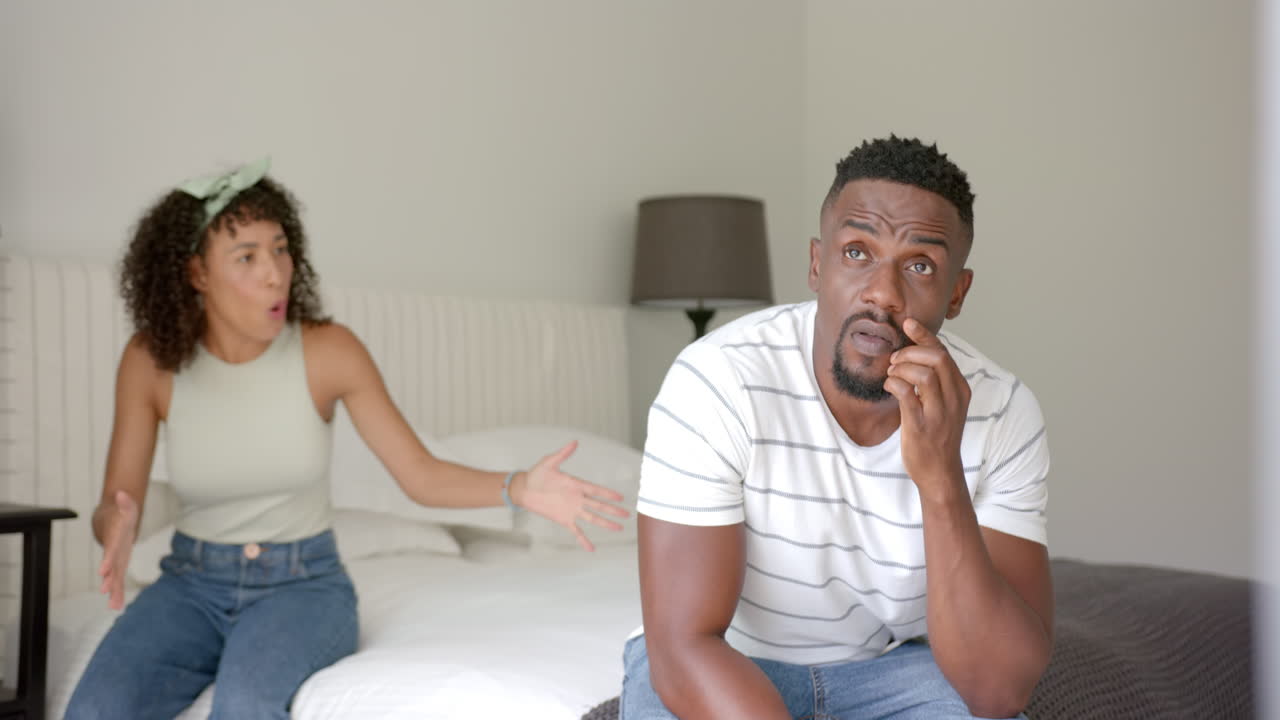 Arguing couple sitting on bed, man rolling eyes and woman gesturing angrily
