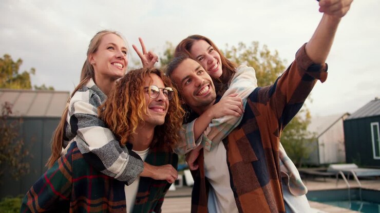 Two girls are sitting on their boyfriends' backs in checkered shirts. A guy with long hair takes a funny selfie near the sunbeds. Rest in the country house