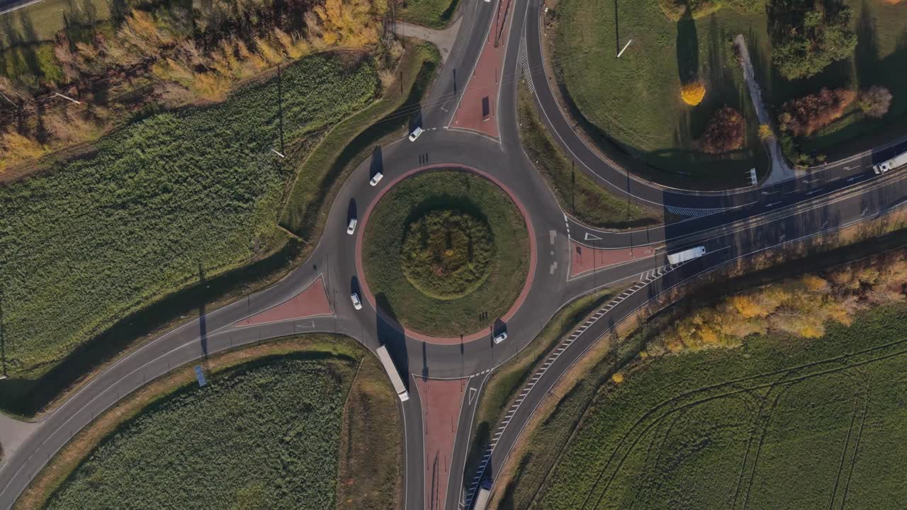 A top-down aerial video of a highway roundabout with trucks and cars moving along the lanes. The circular road is surrounded by green fields and autumn foliage