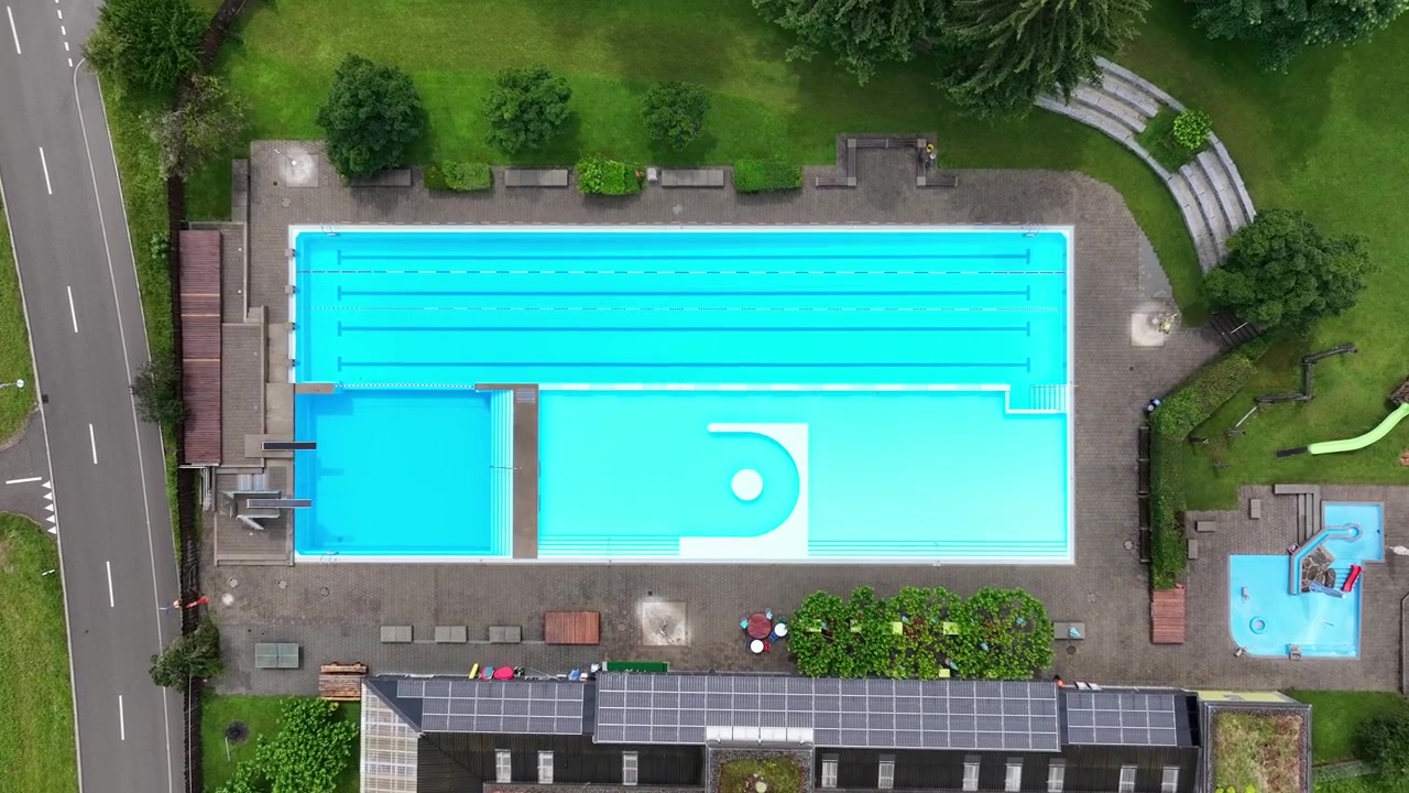 Drone flyver ned over en swimmingpool i Schweiz med svømmebaner og soppebassiner, omgivet af grønne områder.