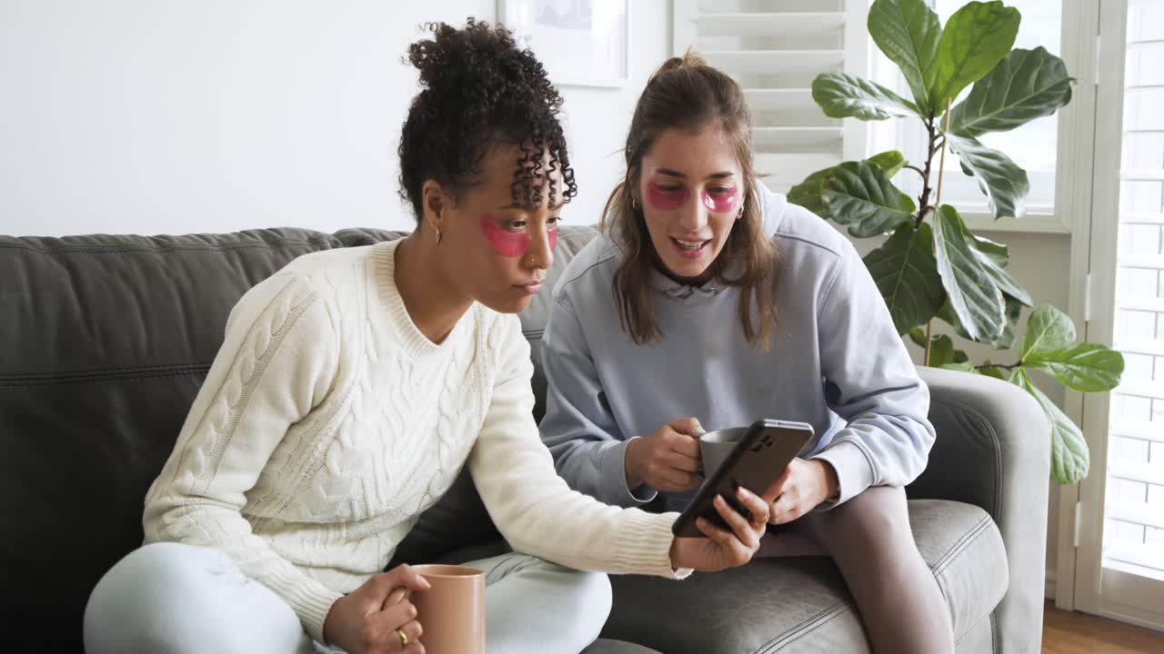 Diverse female friends laughing while swiping tablet on sofa after left friend raising it with mugs