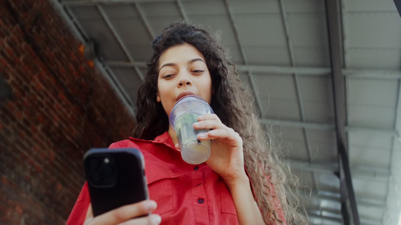 mujer joven bebiendo limonada y usando un teléfono inteligente al aire libre