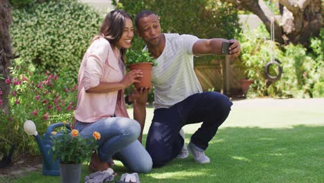 Feliz-Pareja-Birracial-Haciendo-Jardinería,-Tomándose-Selfie-Con-Maceta-De-Flores