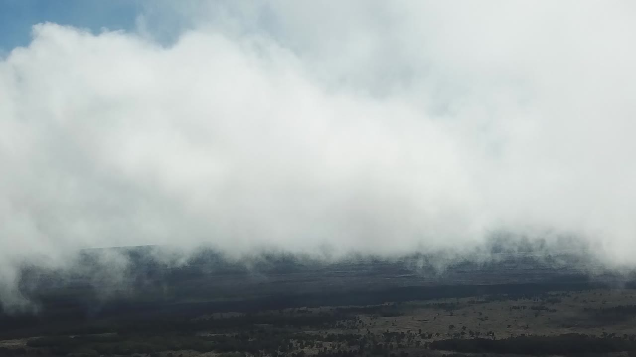 Thick white clouds roll across a dark volcanic slope, partially obscuring the rugged terrain beneath a vivid blue sky, creating a moody natural scene.