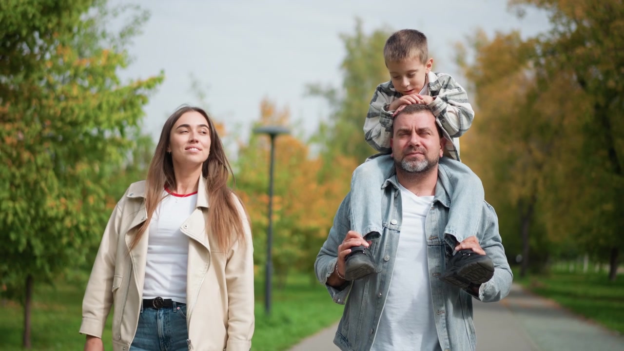 Famiglia che cammina insieme in un parco con l'uomo che porta un bambino sulle spalle e la donna accanto a loro. L'uomo tiene delle scarpe mentre il bambino è seduto in sicurezza; tutti e tre si muovono lungo un sentiero circondato da alberi verdi.