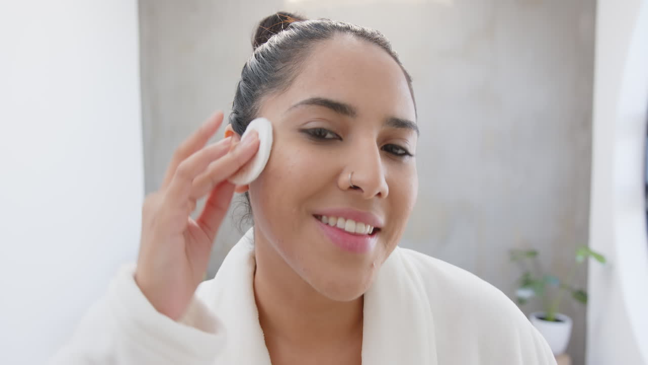 Donna asiatica matura in accappatoio da spa che applica un detergente viso, godendosi la routine di cura della pelle, a casa.