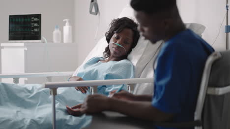 african-american woman resting in hospital bed after surgery talking to young male nurse. portrait of african-american nurse assisting ill female patient lying in bed