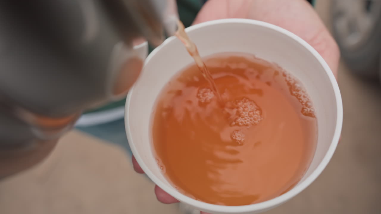 Pouring Tea Into Paper Cup From Thermos Held In Hand On Cold Urban Sidewalk, Steam Rising, Volunteer Serving Warmth And Comfort, Donation Scene Near Parked Cars, Compassionate Community Outreach