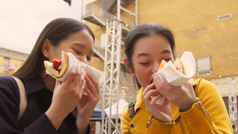 two young female friends eating hot dogs bought at street food market stall