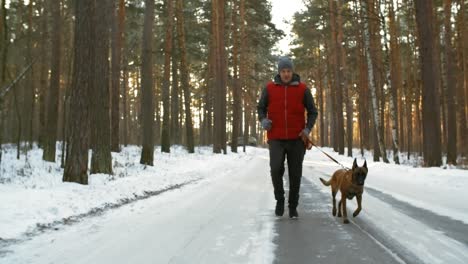 Active-Middle-Aged-Man-Running-On-A-Snowy-Road-In-Forest-With-Belgian-Shepherd-Dog-On-A-Winter-Day