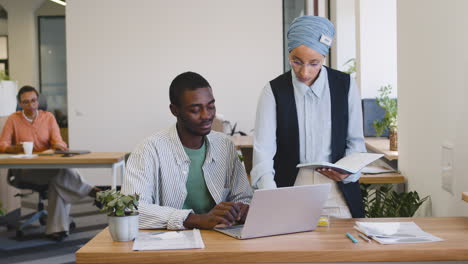 young worker working with laptop sitting at his desk while muslim businesswoman talks to him stand