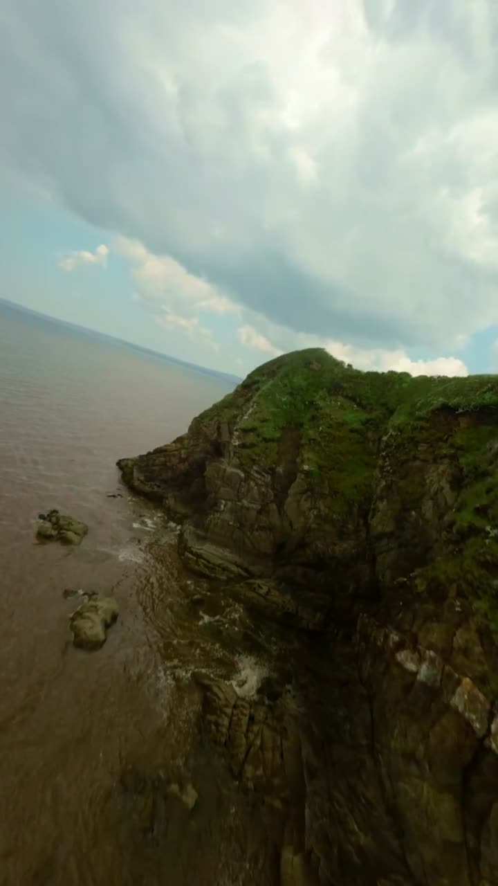 Sobrevoo em baixa altitude com FPV sobre penhascos gramados acima da praia da Bretanha, com terreno acidentado e casas costeiras tranquilas, vertical