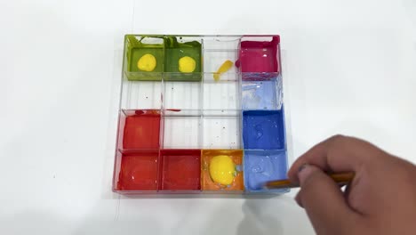 a hand uses a paintbrush to mix blue watercolor paint in a plastic tray, viewed from above on a white background with even lighting
