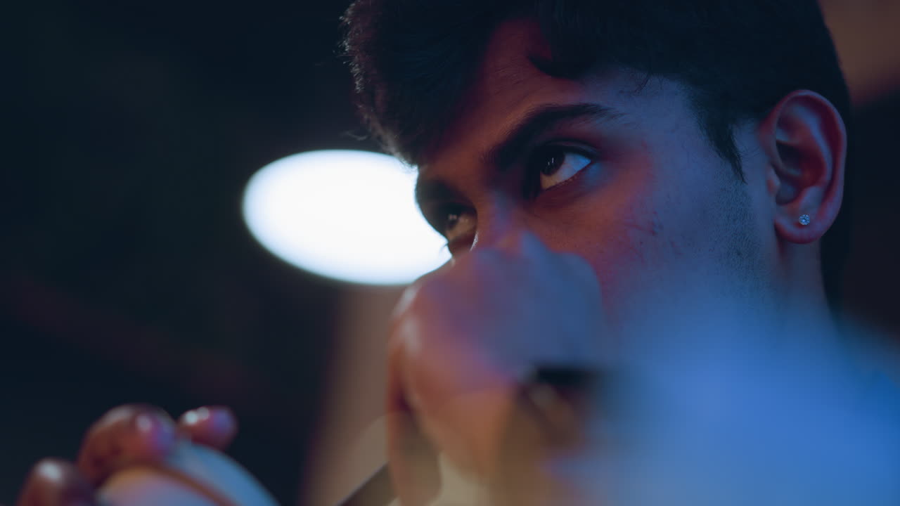 Close-up of young man drinking from takeaway cup through black straw with intense focused gaze, wearing smart shirt and wristwatch, warm ambient lighting and soft blur in background create contemplative atmosphere