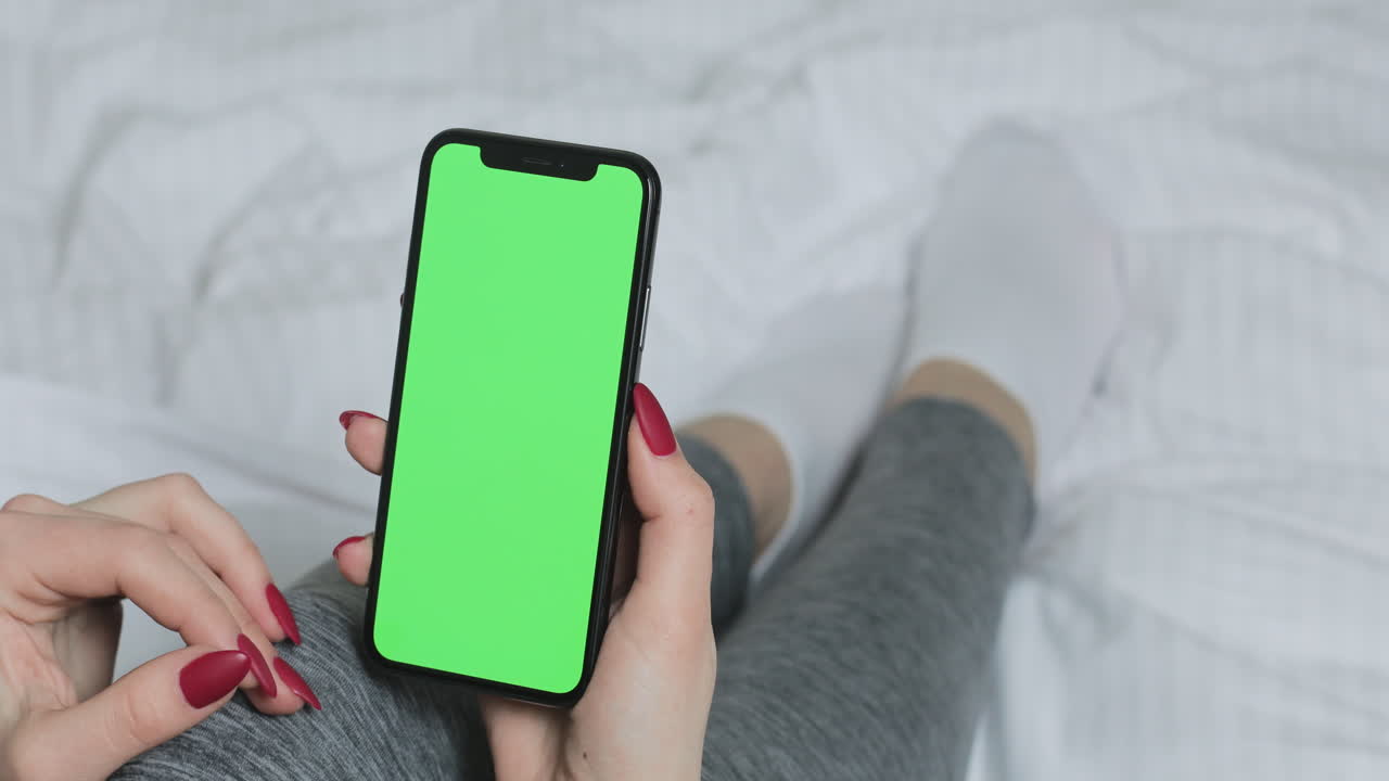 Woman using smartphone with green screen on bed