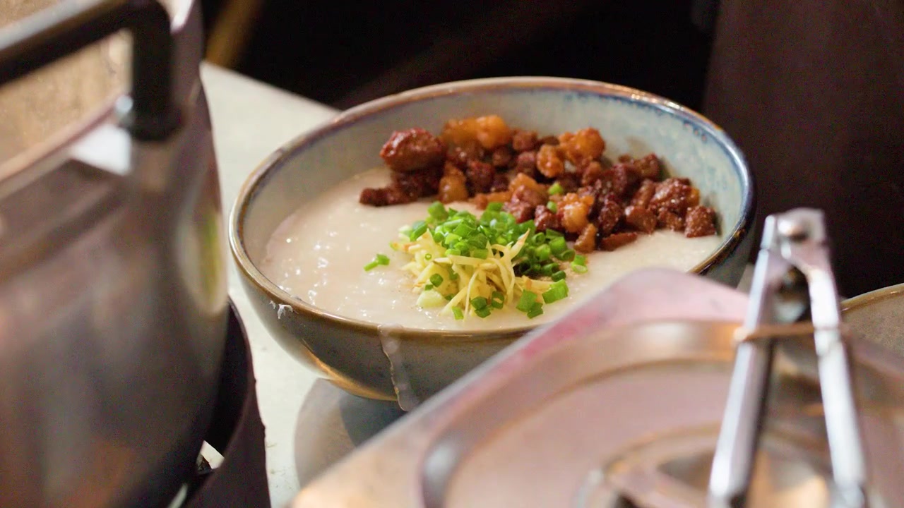 Close-up seorang koki menambahkan daun bawang segar ke dalam semangkuk bubur gurih dengan daging babi