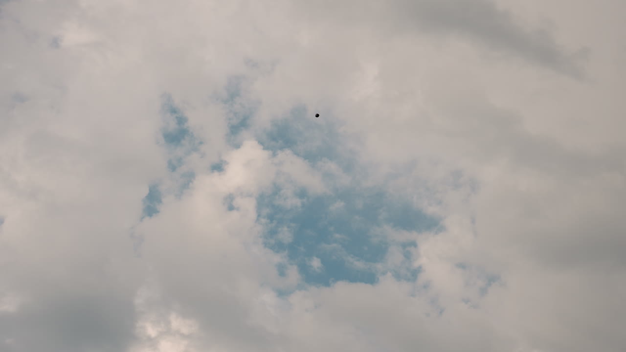 solitary black balloon drifting into vast cloudy sky over open field, floating slowly upward against backdrop of soft white clouds and pale blue hues