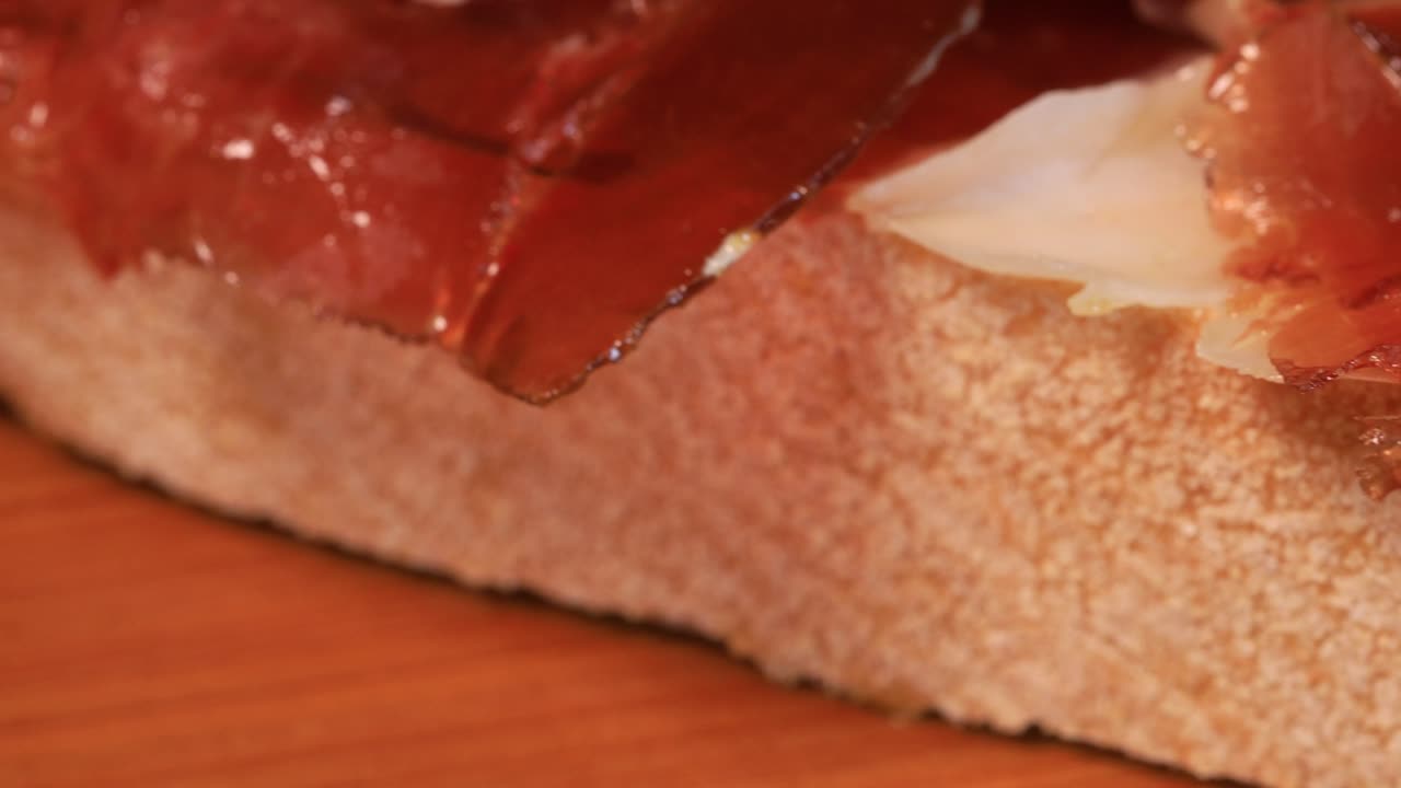 Close up view of oily high quality spanish serrano ham with bread rotating on wooden board, macro shot from above