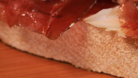 close up view of oily high quality spanish serrano ham with bread rotating on wooden board, macro shot from above